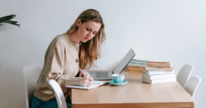 (Focused woman writing in a notebook with a laptop and books nearby, representing personal financial planning and budgeting at home.)