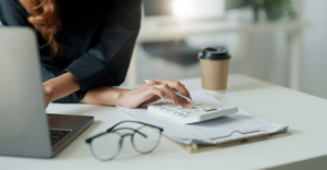 A person using a calculator at a desk with financial documents, a laptop, glasses, and a coffee cup.
