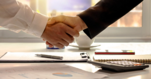 Two people shaking hands over a desk with documents, a pen, a notebook, a calculator, and a coffee cup.