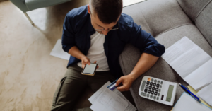 A man sitting on the floor next to a couch, holding a phone and a credit card, surrounded by papers and a calculator.