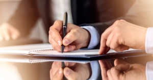 Close-up of two people reviewing and signing documents on a reflective table, with one hand holding a pen and the other pointing at the paper.