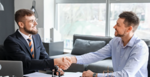 Two men sitting across from each other at a desk, smiling and shaking hands in a bright office setting.