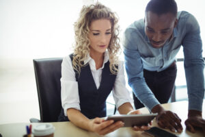 Serious focused colleagues using tablet together, looking and pointing at gadget screen while sitting at table in office. Communication or teamwork concept